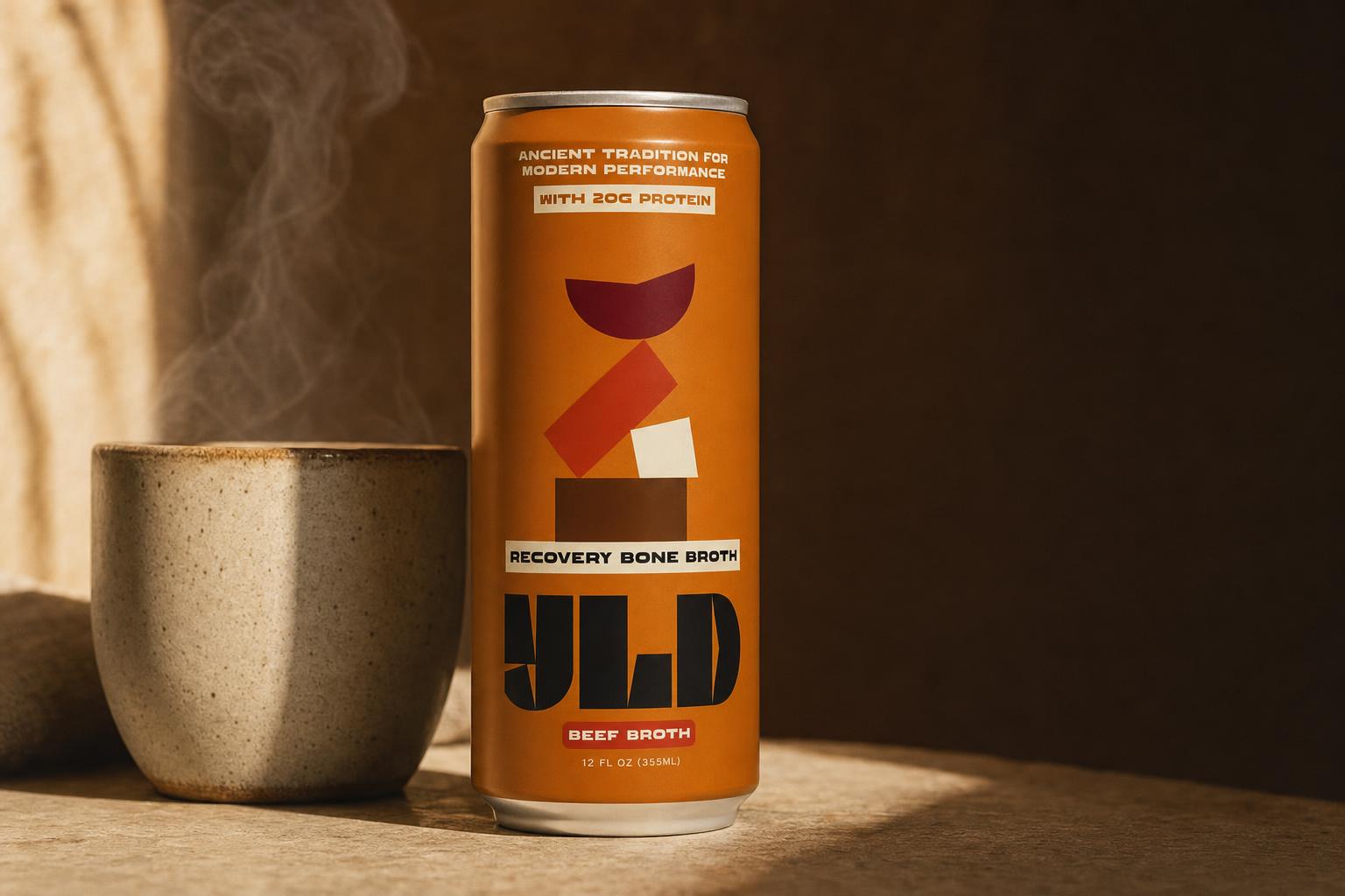 A YLD beef bone broth can beside a steaming ceramic cup in warm light
